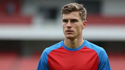 A determined, 28-year-old Hvidovre midfielder, with a strong build and focused expression, wears a plain red and sky blue...